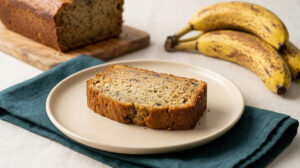 Une tranche de banana bread sur une assiette beige, posée sur une serviette bleu canard avec des bananes en arrière-plan.